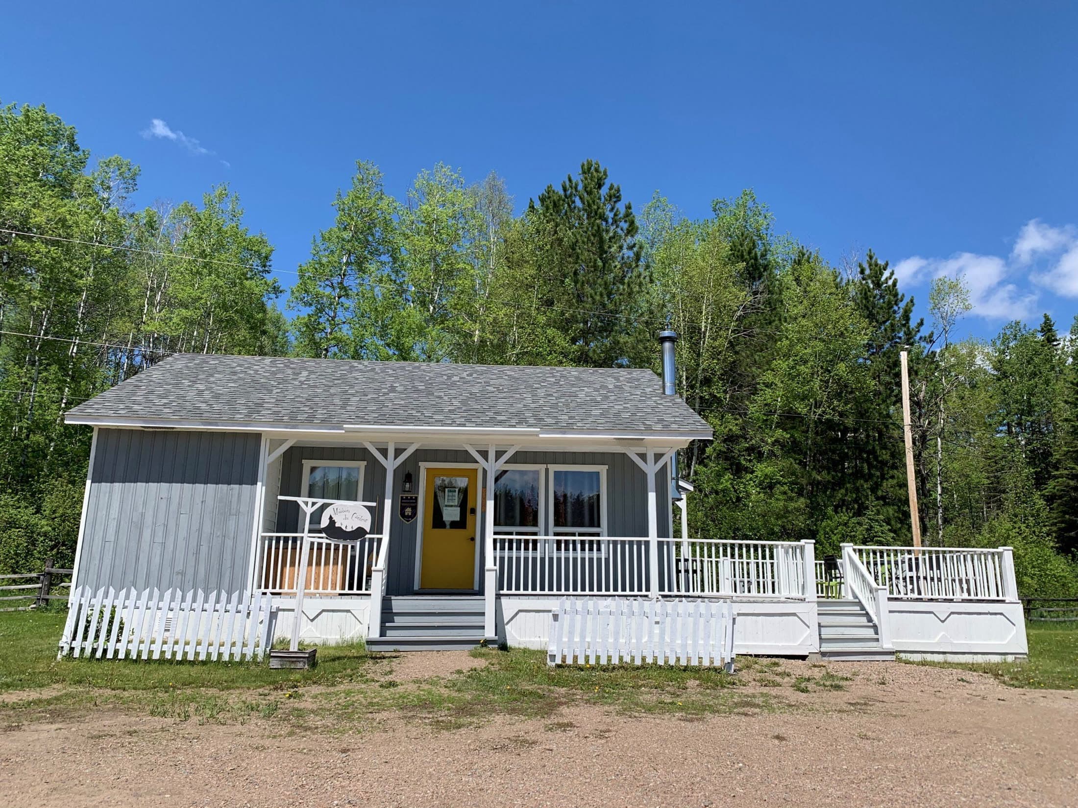 La Maison du Conteur, chalet gris à porte jaune, galerie couverte et clôture blanche, sous un ciel bleu.