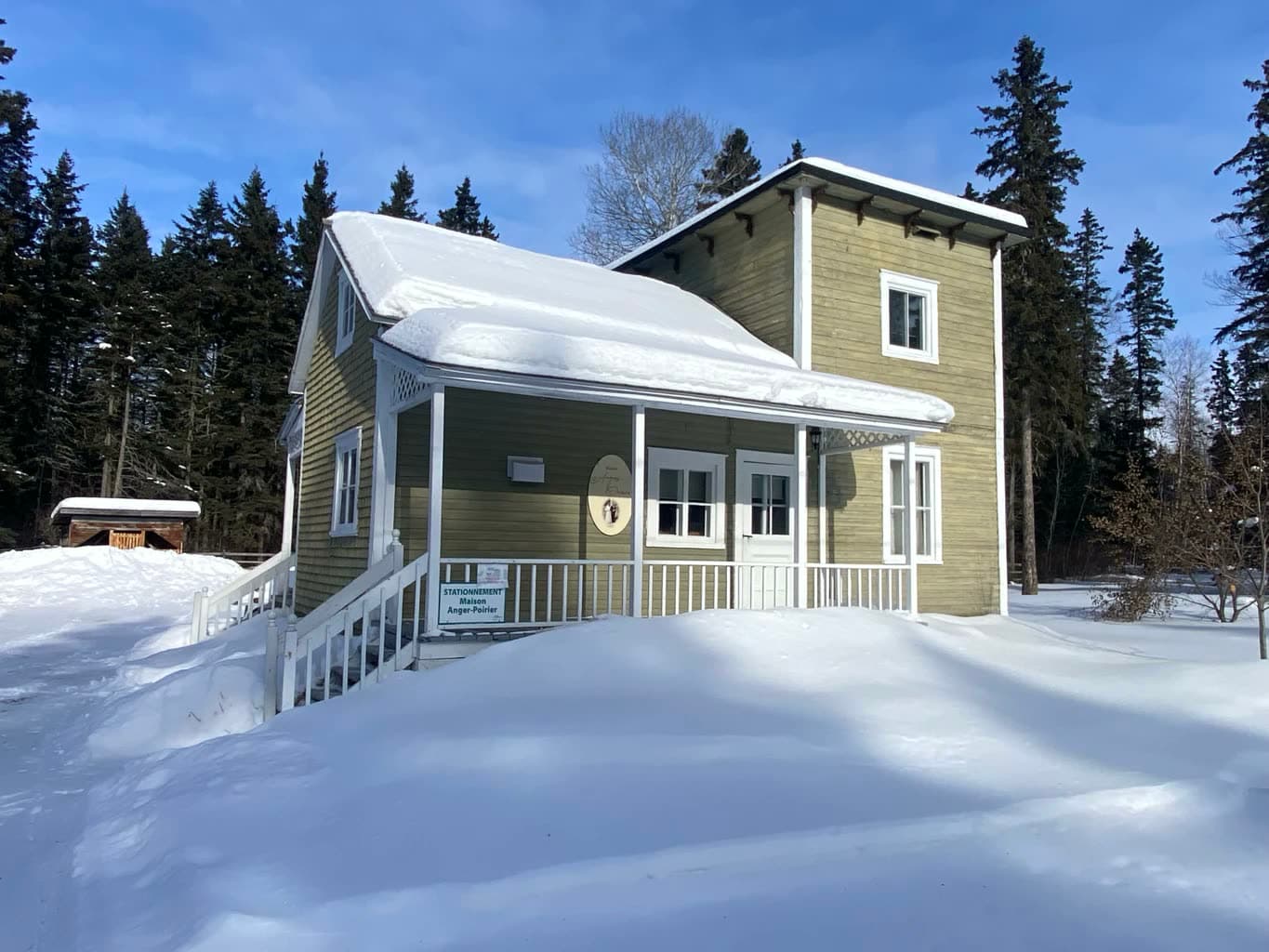 La maison sous la neige, sa galerie couverte et les sapins derrière.